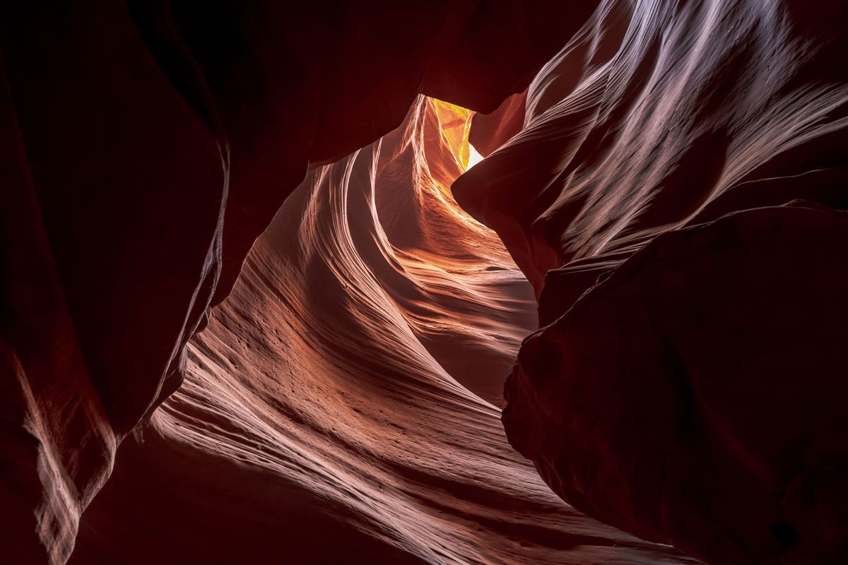 Looking up through a narrow slot canyon with curving red and orange sandstone walls and a bright glowing opening at the top.