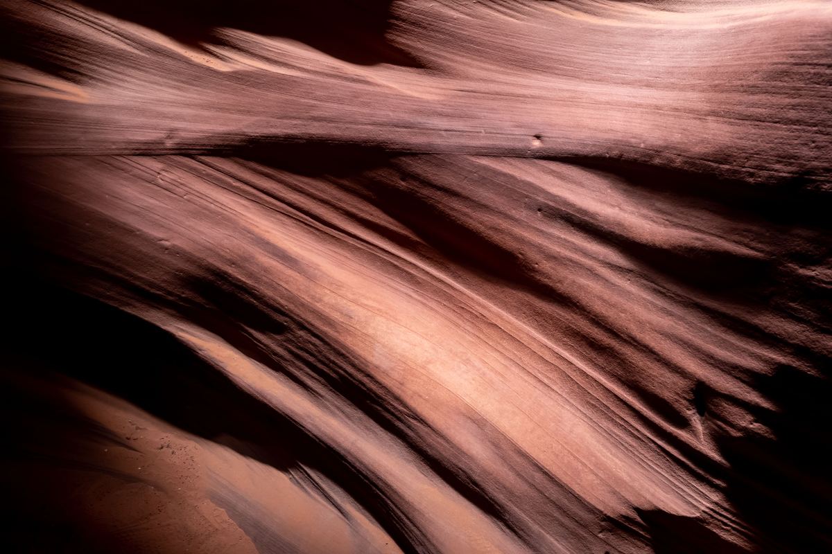 Close view of smooth, curving sandstone walls with flowing layered lines in warm pink and brown tones, fading into dark shadow.